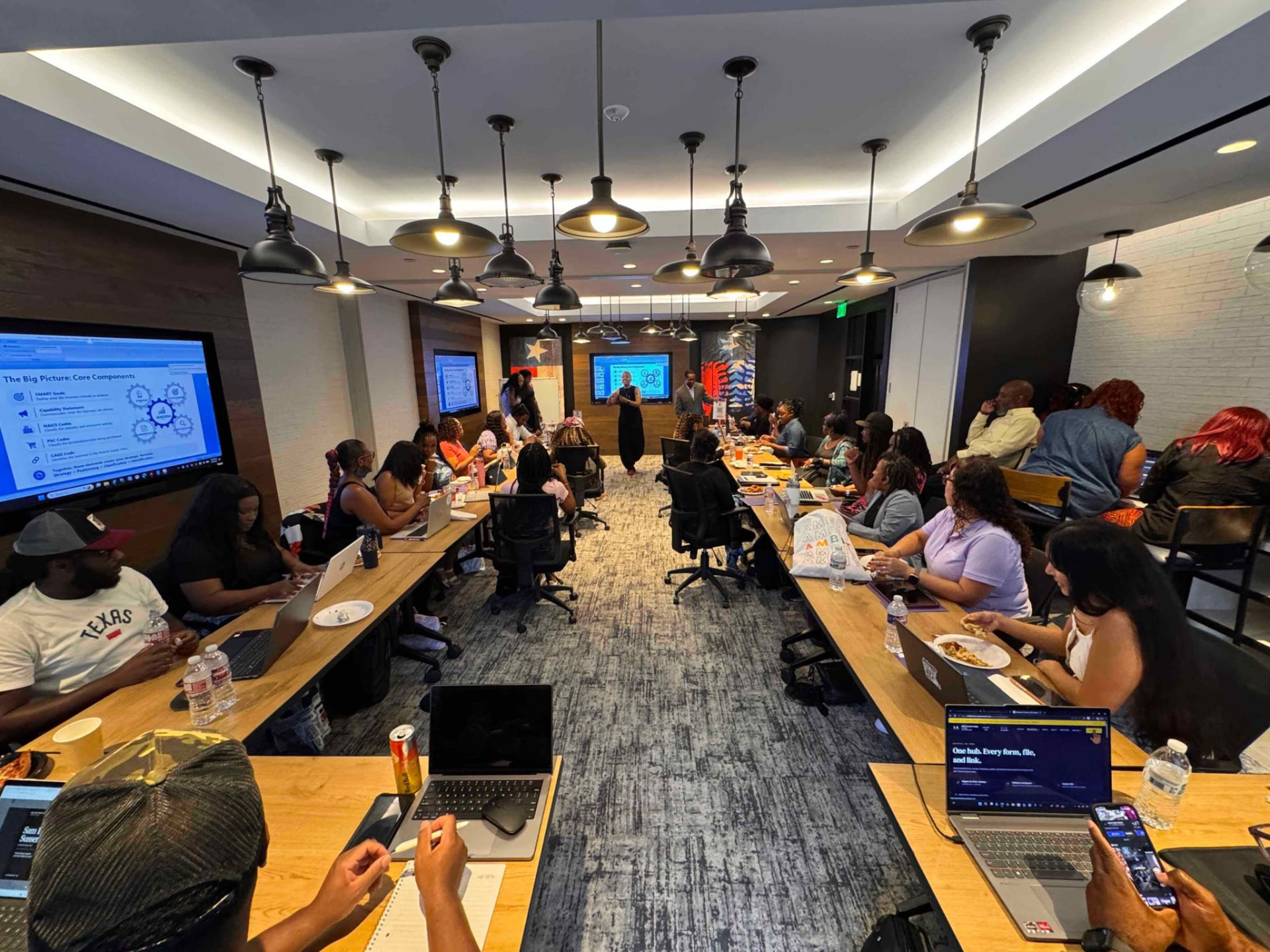 B.I.D. Academy participants seated along two long tables under pendant lights while a slide on core business components is projected.