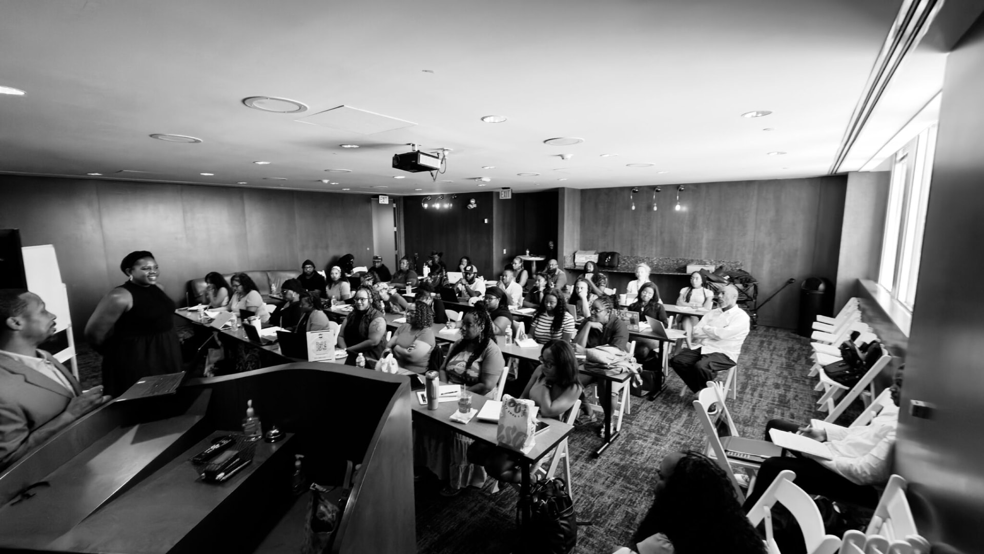Wide black-and-white view of a B.I.D. Academy session, the facilitator at the front and every table filled.