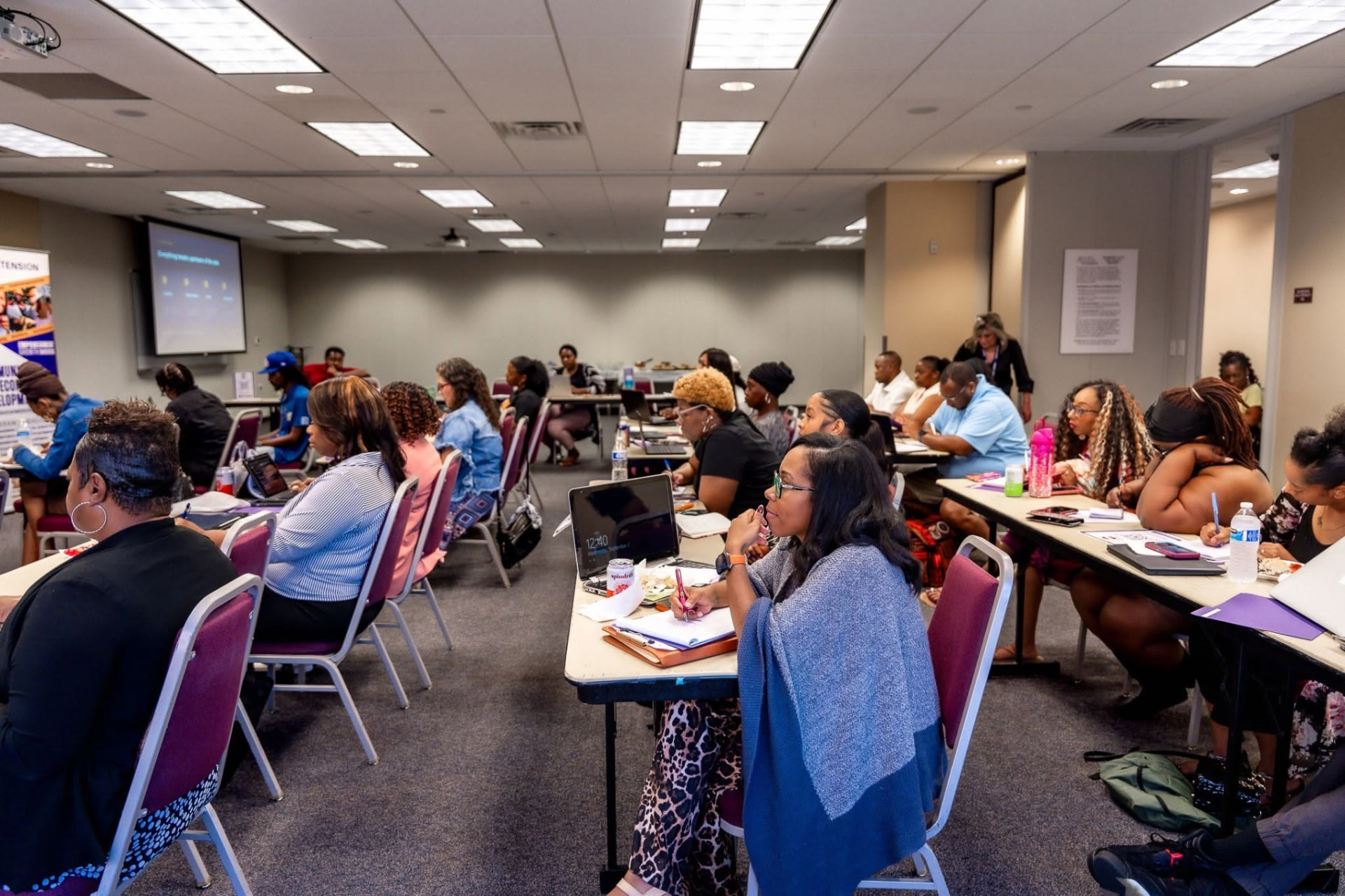 A wide view of a B.I.D. Academy session in progress, participants at tables with laptops and a slide on screen.