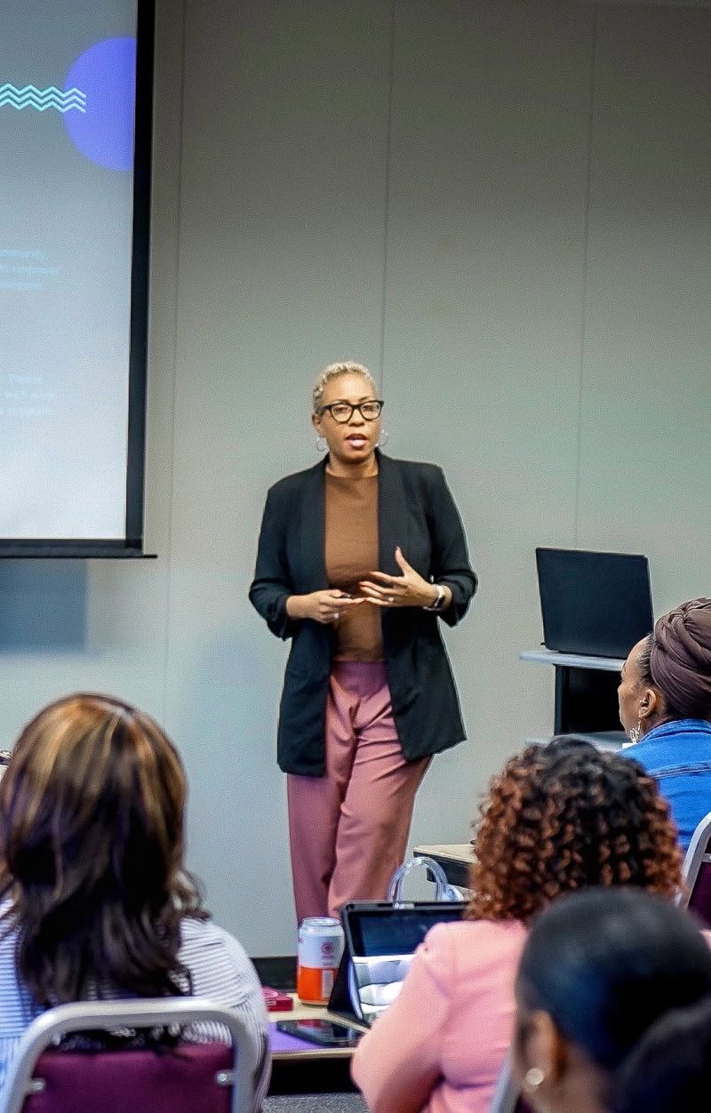 A facilitator in a black blazer speaking to seated B.I.D. Academy participants beside a projection screen.